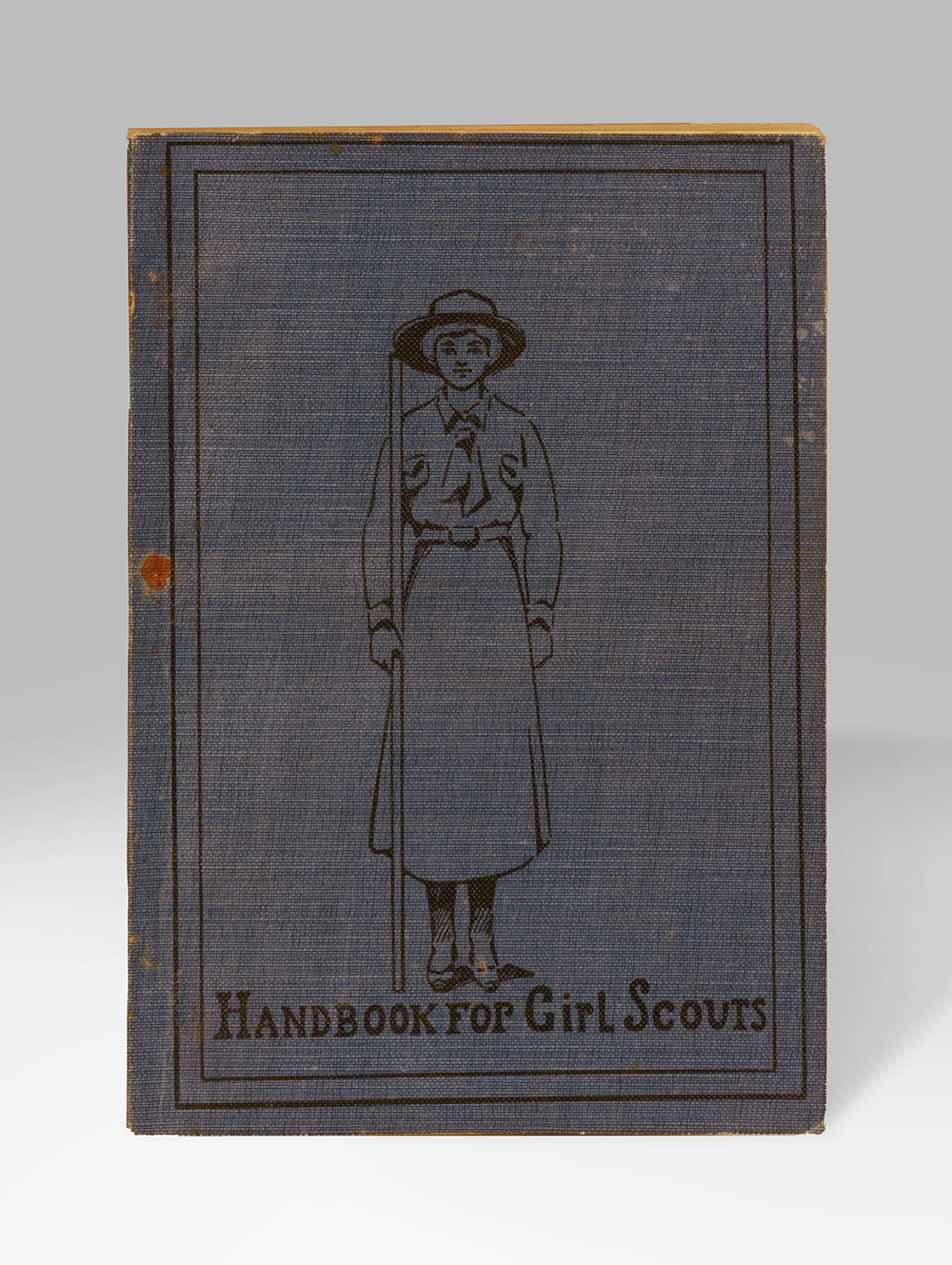 Livre fermé avec une couverture gris foncé illustré d'un dessin au trait d'une jeune fille portant un chapeau, une chemise et une jupe uniformes. Texte : « Manuel pour les éclaireuses »