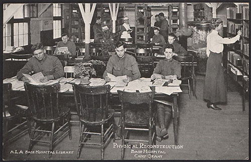 Black and white photograph of a room lined with bookshelves and filled with tables and chairs. Several white male soldiers sit at the table in the foreground reading pamphlets or papers. A white woman in a simple blouse and long skirt reaches for books in one of the side bookshelves. More men and women mill around in the background holding books or are seated at background tables reading.