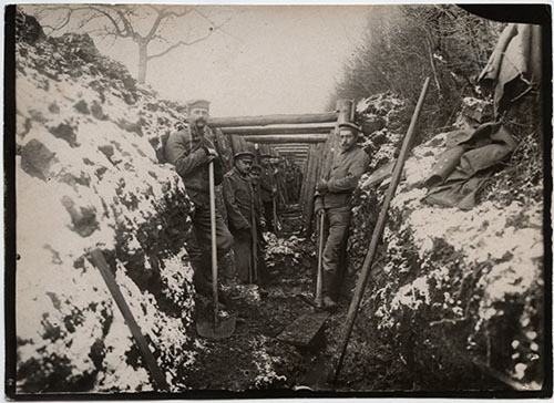 Black and white photo of a group of soldiers posed facing the viewer, leaning against the earth walls of a trench holding shovels and pickaxes.