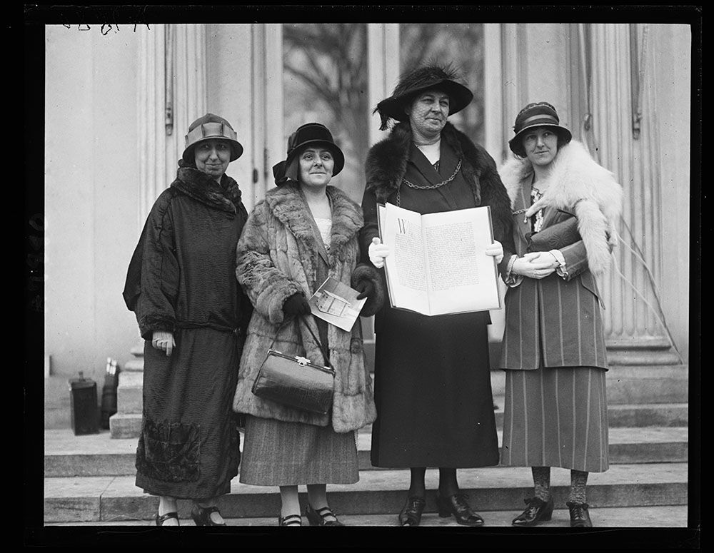 Black and white photograph of four white women dressed in fine traveling dresses, coats and hats posing outside with one woman holding open a large folio-like object with writing on the pages.