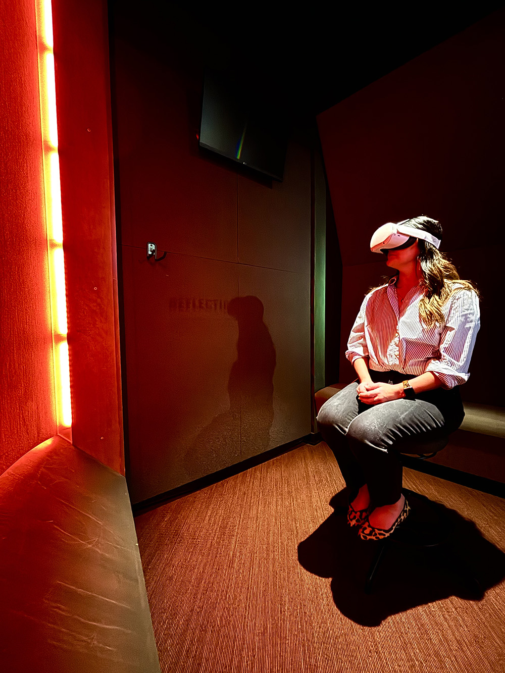Modern photograph of an enclosed alcove in a museum gallery. A white woman with long brown hair wearing jeans and a button-down sits on a comfortable bench, wearing a large VR headset over her eyes.