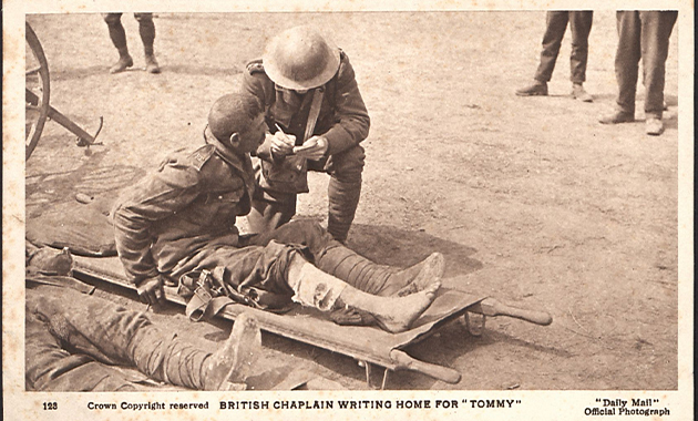 Sepia photograph of two soldiers in combat uniforms outside somewhere. One man is sitting up on a stretcher that is on the ground. The other man is kneeling next to him with pen and notebook, which the two of them are conferring over.