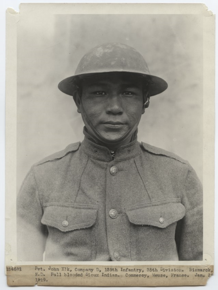Black and white portrait photograph of a young Native American man in WWI U.S. military uniform and metal doughboy helmet.
