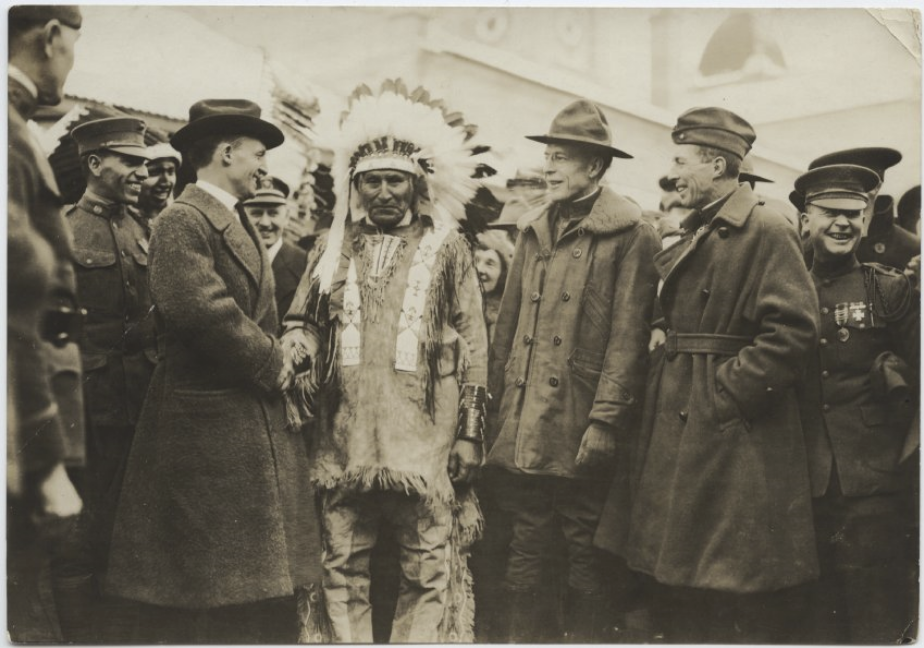 Black and white photograph of a group of men talking jovially. In the center, one man is wearing ceremonial buckskins and a feathered headdress. The rest are in American/European-style coats and hats.