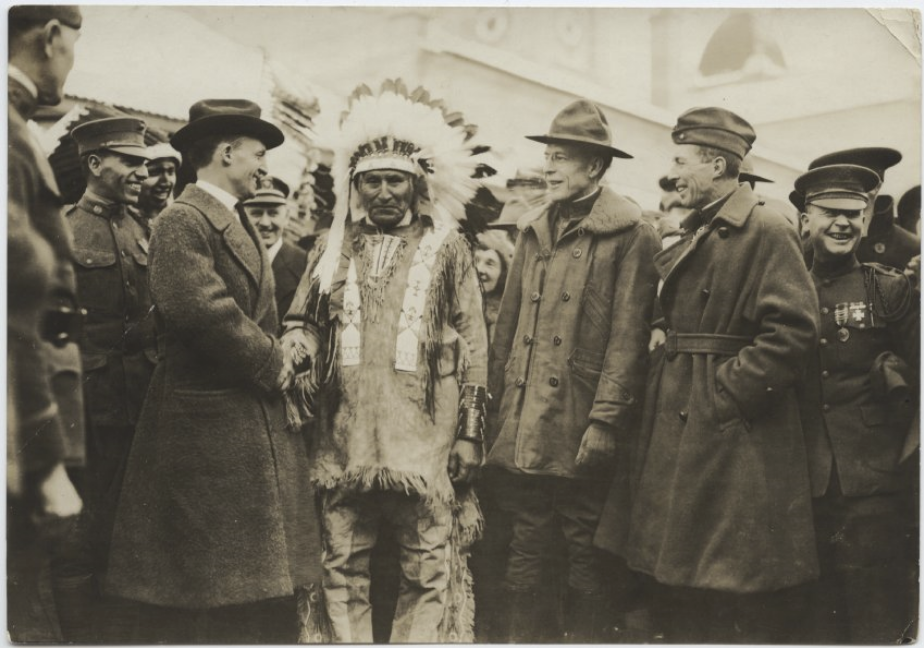 Photographie en noir et blanc d’un groupe d’hommes discutant jovialement. Au centre, un homme porte des peaux de daim de cérémonie et une coiffe à plumes. Les autres portent des manteaux et des chapeaux de style américain/européen.