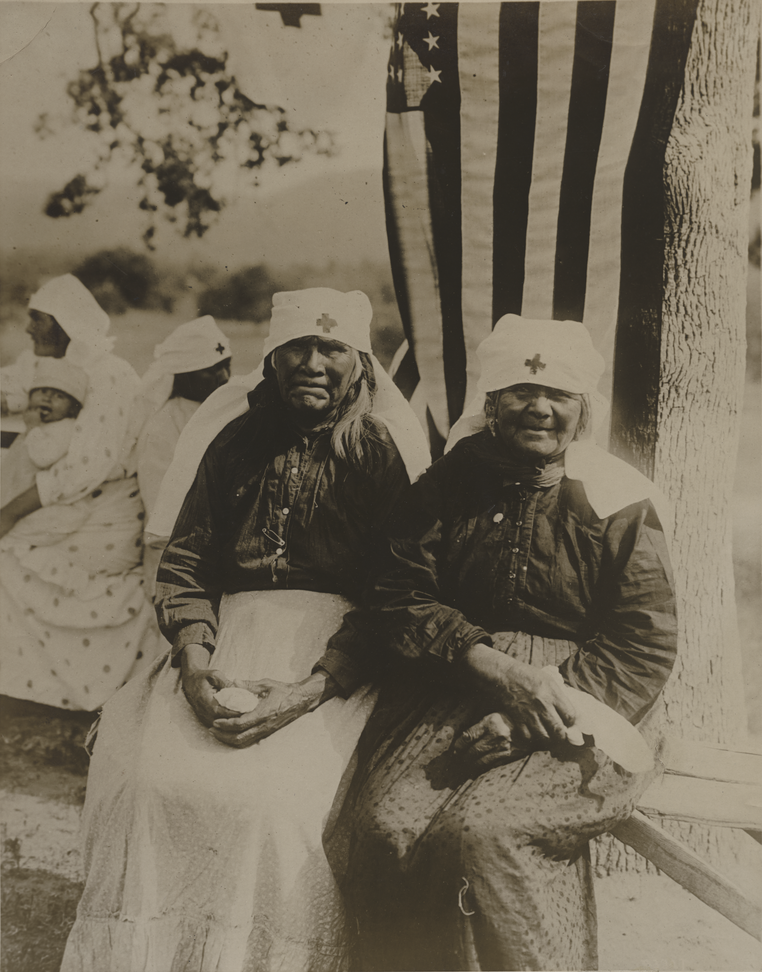 Black and white photo of two elderly Native American women in Red Cross uniforms and headdresses, seated under a tree from which a U.S. flag has been draped.