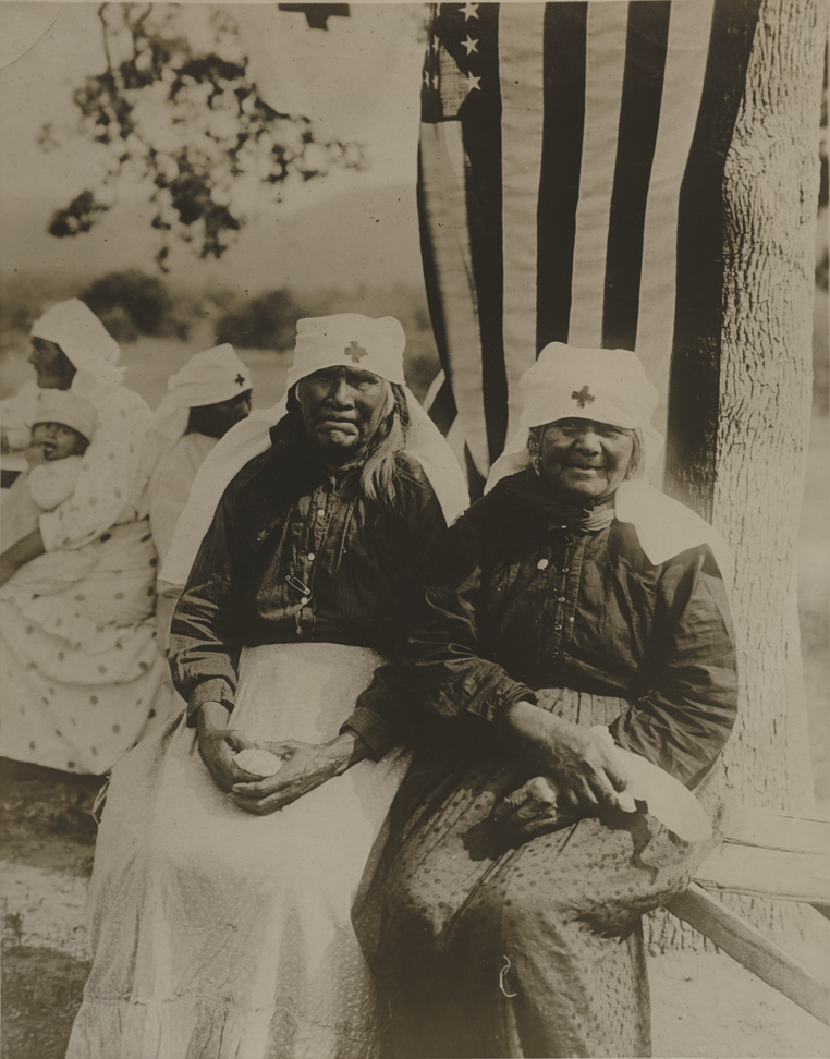 Photo en noir et blanc de deux femmes amérindiennes âgées portant des uniformes et des coiffes de la Croix-Rouge, assises sous un arbre sur lequel un drapeau américain a été drapé.