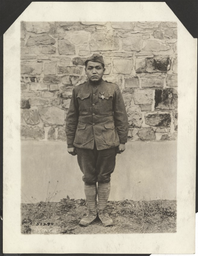 Black and white portrait photograph of a young Native American man in WWI military uniform standing at attention in front of a stone wall.