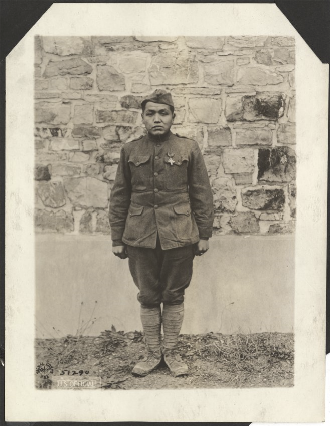 Photographie portrait en noir et blanc d'un jeune amérindien en uniforme militaire de la Première Guerre mondiale, au garde-à-vous devant un mur de pierre.