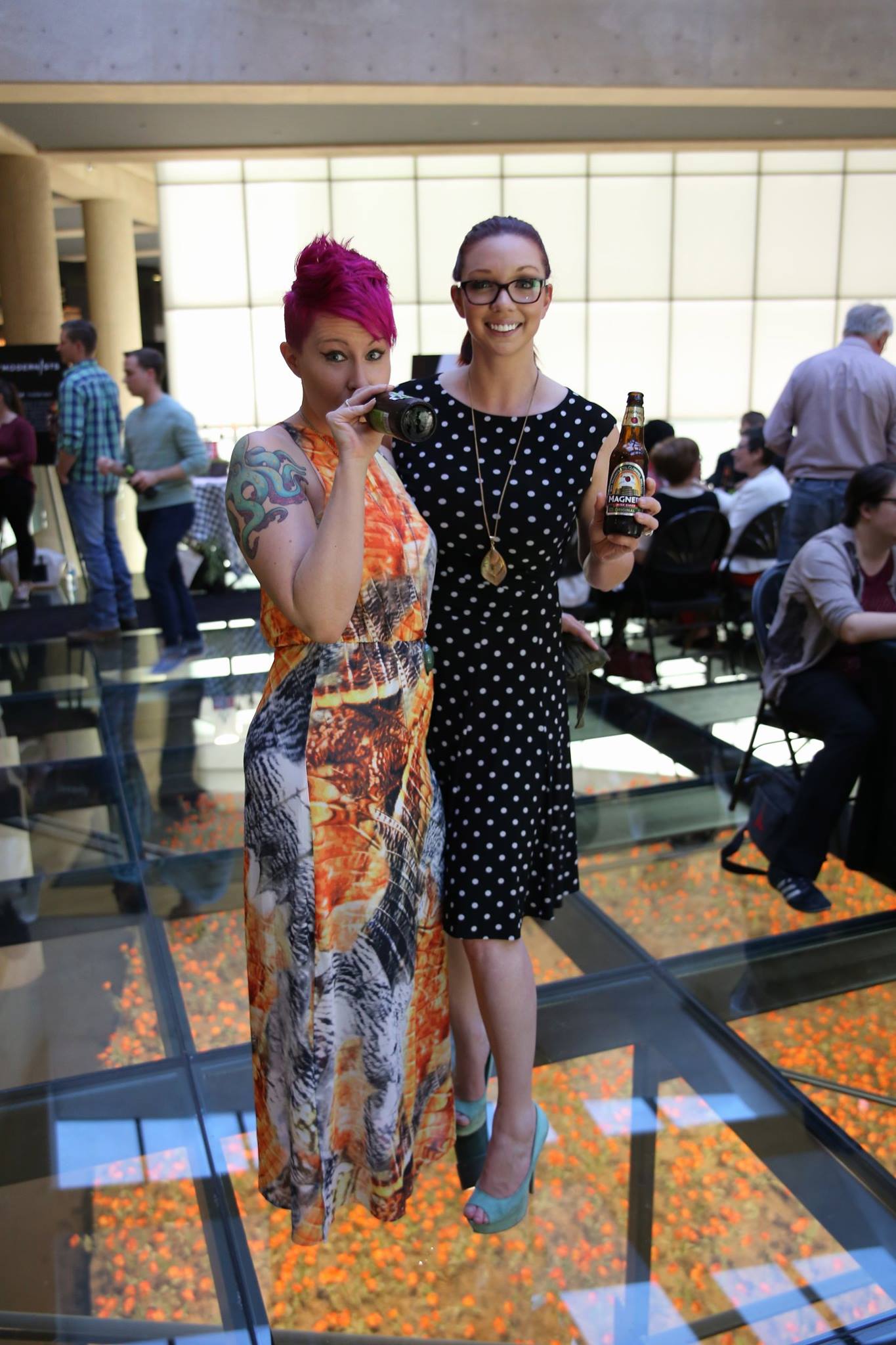 Modern photograph of two young white women in cocktail attire posing on the Glass Bridge. One woman has spiky pink hair and is drinking from a glass bottle of cider, while the other is smiling and holding her bottle up for the viewer to see.