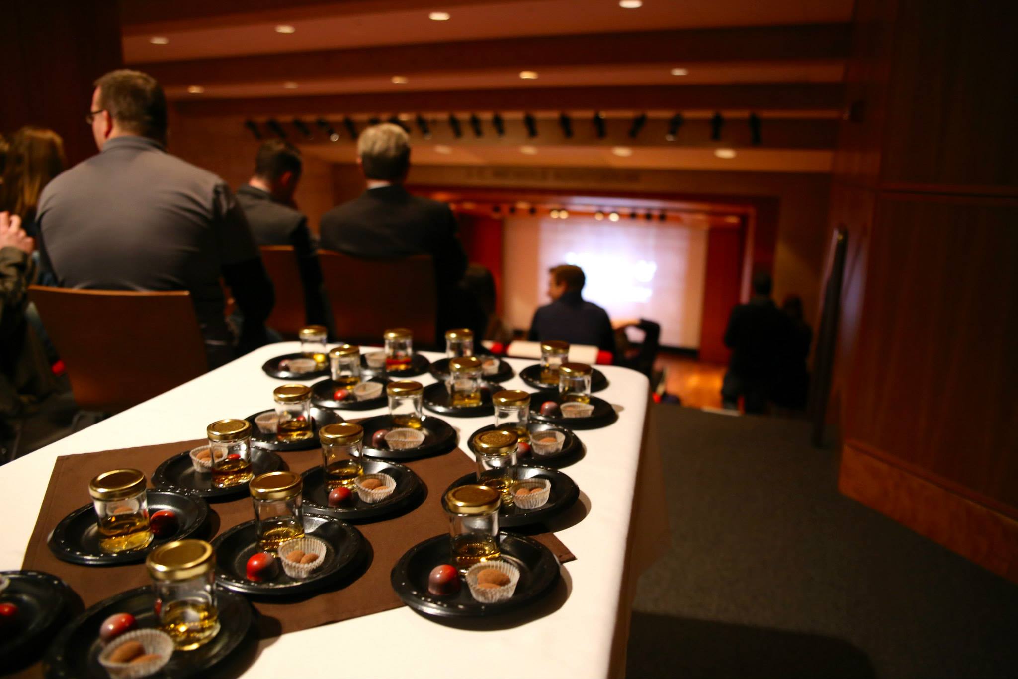 Photographie moderne d'une table dressée au fond de l'Auditorium, remplie de monde. La table est remplie de nombreuses petites assiettes contenant chacune des shots de whisky et des chocolats.