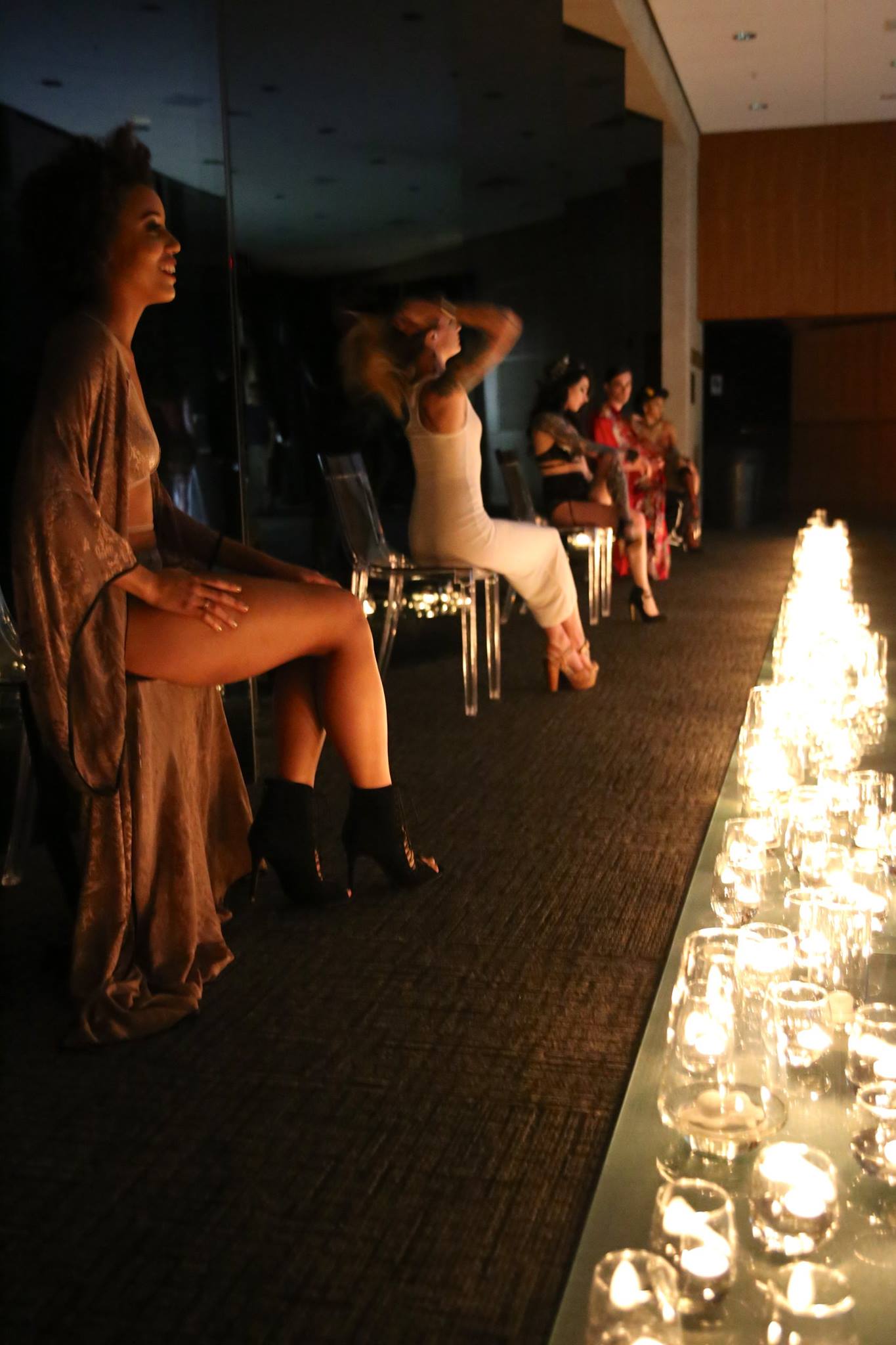 Modern photograph of several young, slim models of different skin tones and genders, seated in a row in the Auditorium Lobby, wearing vintage and vintage-inspired lingerie and undergarments. They are dimly illuminated by dozens of tea candles in glass cups and vases.