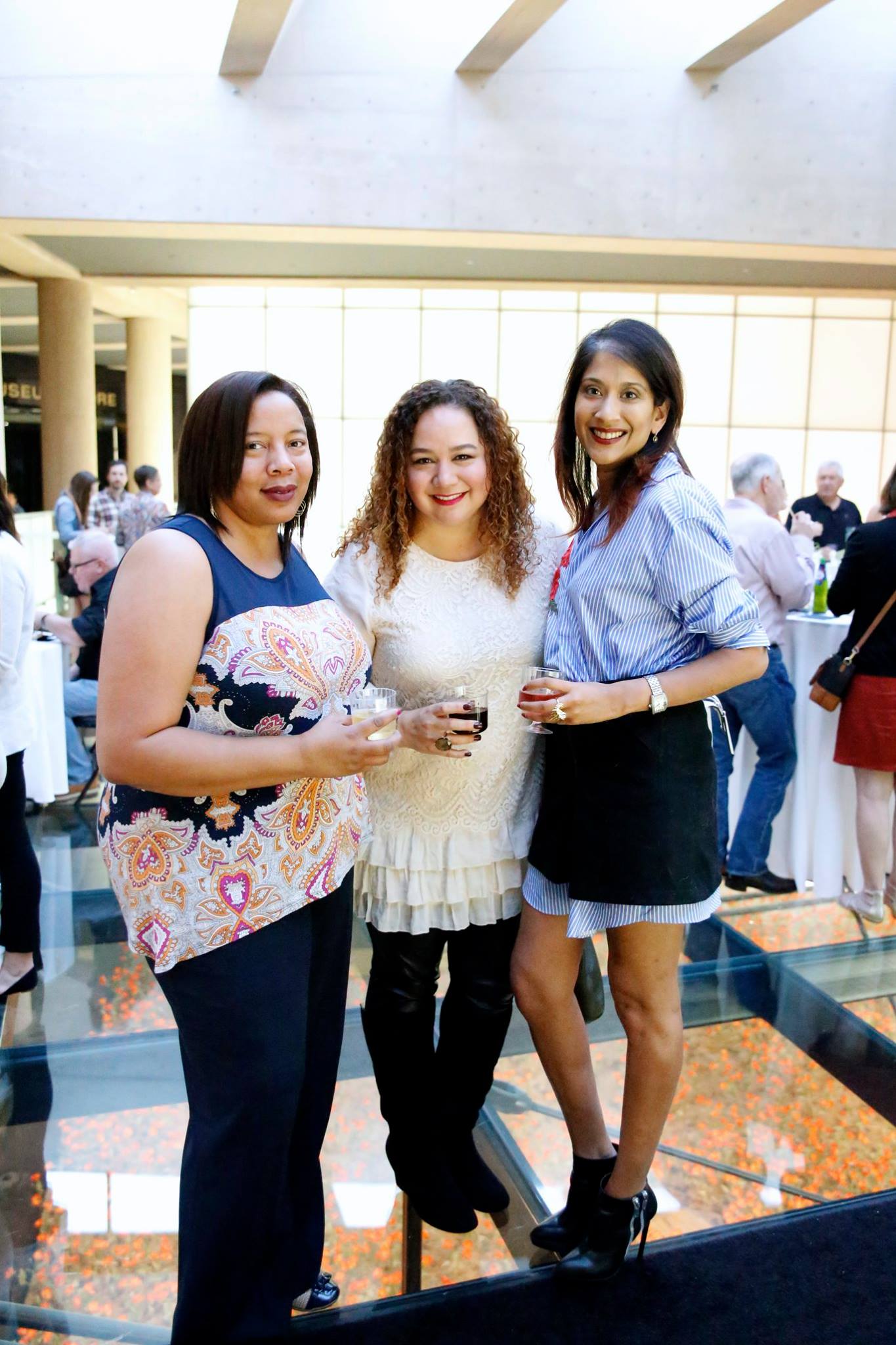 Modern photo of three smiling, light brown-skinned women in casual cocktail outfits posing on the Glass Bridge with glasses of wine in their hands.