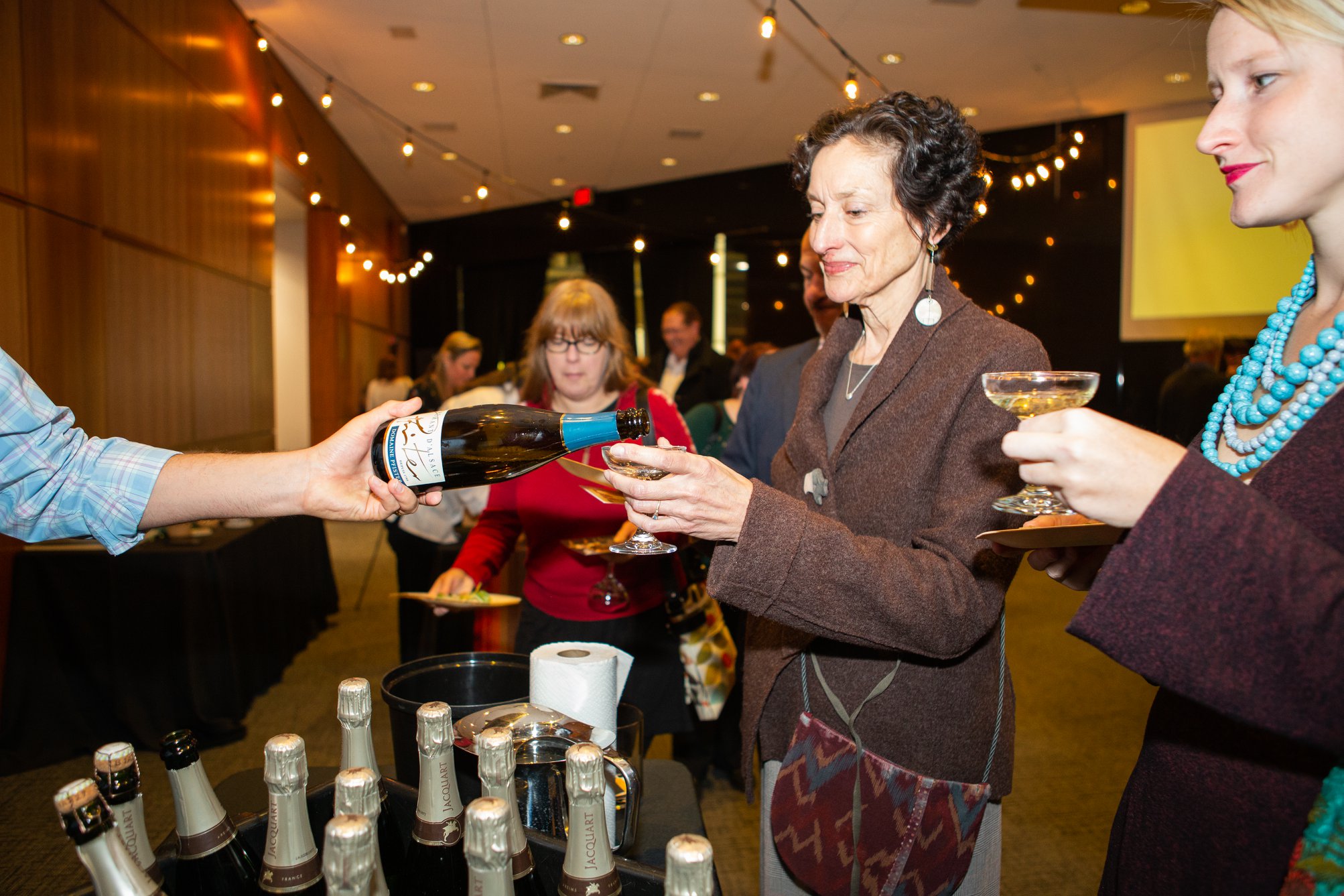 Modern photograph of a middle-aged white woman in a stylish brown blazer holding a champagne coupe glass up for someone out of the frame who is pouring champagne from a bottle into the glass. Several other women guests stand in line waiting for their turn.