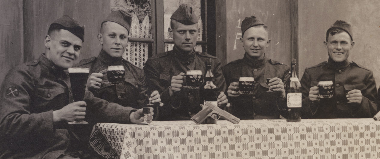 Black wnd white photograph of five young white men in WWI military uniform sitting in a row at a table, toasting the viewer with large pint glasses of beer.