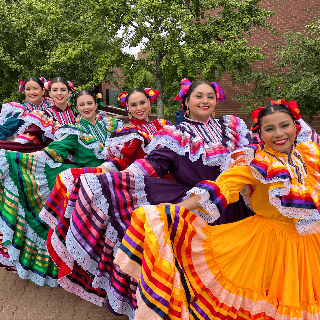 Modern photograph of six dancers dressed in extravagantly colorful traditional Mexican dresses, smiling at the viewer and fanning their skirts out to show off the patterns.