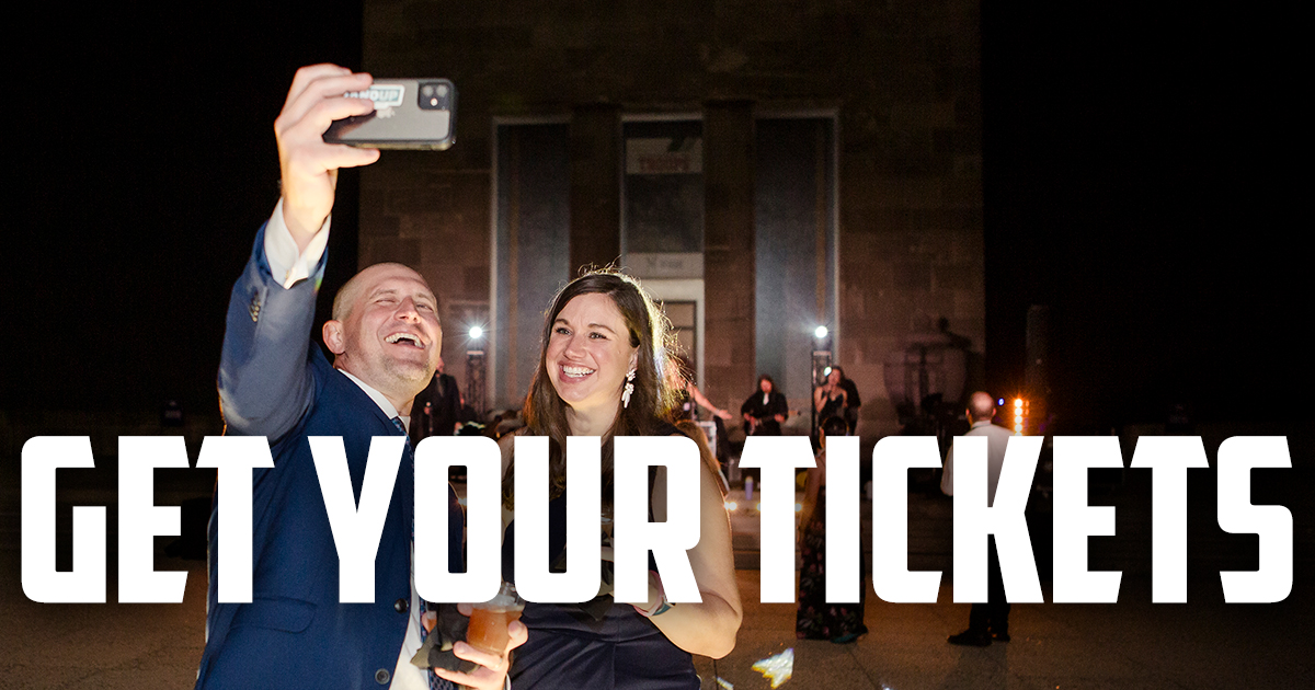 Modern photo of the Memorial Courtyard at night, lit up with stage lights and accent lights. A white man in a blue suit takes a selfie with a white woman in a dark blue cocktail dress, both of them smiling and holding drinks. Text: 'Get Your Tickets'