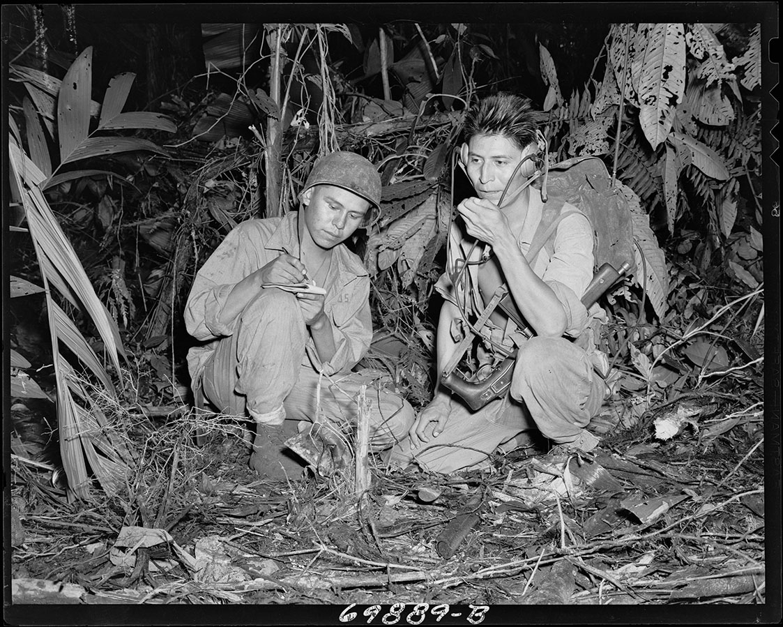 Photographie en noir et blanc de deux hommes amérindiens en uniforme de combat de la Seconde Guerre mondiale, agenouillés dans une clairière de la jungle. L’un parle dans une radio portable tandis que l’autre écrit sur un bloc-notes.