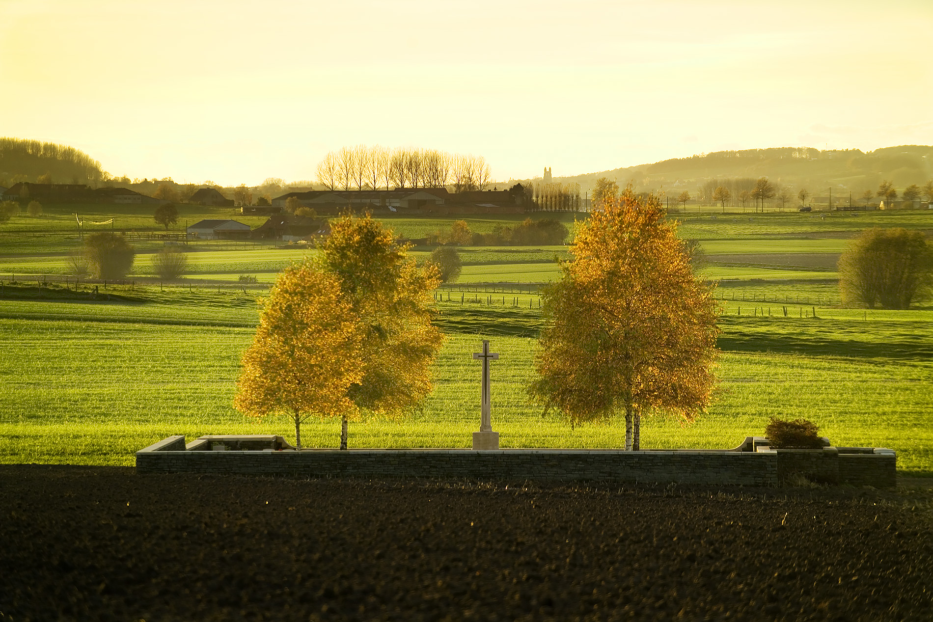 Modern photograph of wide green fields dotted with autumnal trees. A tall cross memorial is in the center foreground.