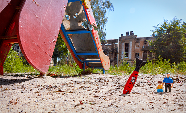 Two plastic dolls resembling a father and son stand in the sand near a rusted playground slide. Nearby, sticking out of the sand nose down, is a toy missile painted to look like a red fish with a black tail.