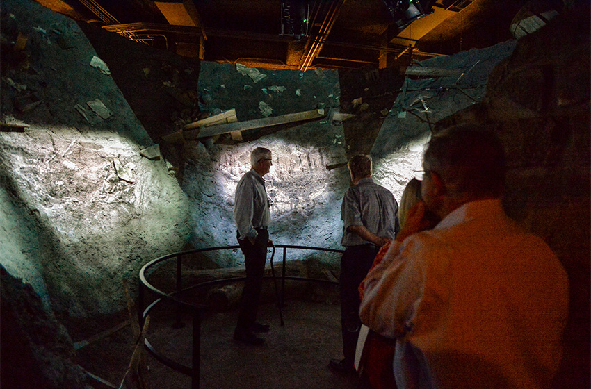 Modern photograph of a museum exhibit made to look like the inside of a bomb crater scattered with the rubble of trees and a house. Black and white film footage is projected onto the crater walls as museum guests look on.