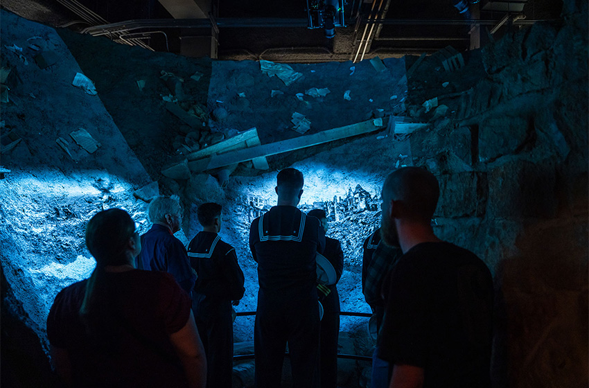 Modern photograph of a museum exhibit made to look like the inside of a bomb crater scattered with the rubble of trees and a house. Black and white film footage is projected onto the crater walls as museum guests look on.