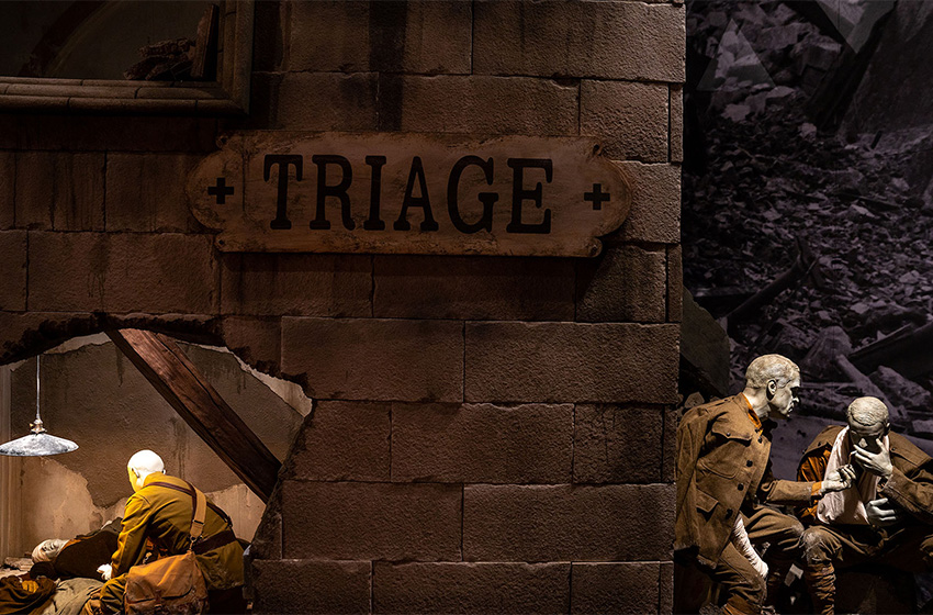 Modern photograph of a museum exhibit made to look like the exterior and interior of a bombed-out stone building. A large sign that reads 'TRIAGE' is hung on the wall. Two soldier mannequins sit 'outside' the building, smoking. On the other side of the building wall, a medic treats wounded soldiers on stretchers.