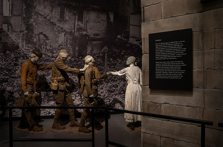 Modern photograph of a scene featuring three soldier mannequins with bandages wrapped around their heads holding on to each other's shoulders as a female nurse guides them toward the entrance of a bombed-out church.