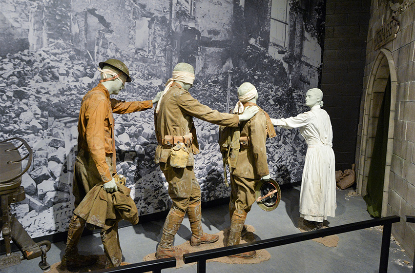 Modern photograph of a scene featuring three soldier mannequins with bandages wrapped around their heads holding on to each other's shoulders as a female nurse guides them toward the entrance of a bombed-out church.