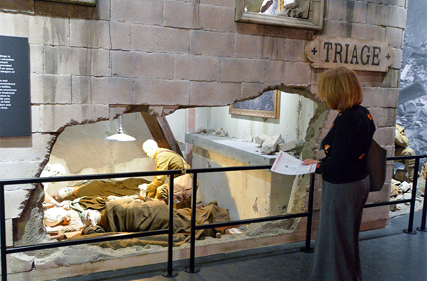 Modern photograph of a museum exhibit design to look like a bombed-out stone building. A museum guest looks through a cutaway wall that reveals a scene with mannequins of injured soldiers lying on pallets while medics and nurses treat them.