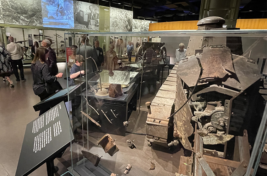 Modern photograph of a museum exhibit of a small WWI tank. Between the viewer and the tank, there is a glass case displaying shrapnel collected from the tank.