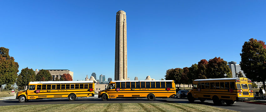 Modern photograph of three yellow school buses lined up in the parking lot in front of the Liberty Memorial Tower against a clear blue sky.