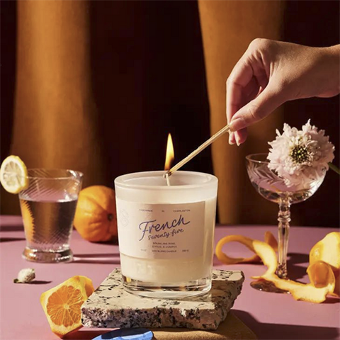 A hand lighting a candle in a cream-colored candle jar, on a pink table surrounded by cocktail glasses, oranges and orange peels