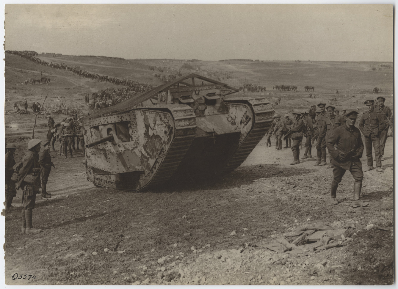 Black and white photo of a small WWI tank rolling through a valley surrounded by soldiers marching beside and behind it.