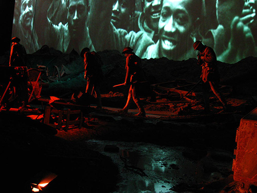 Modern photograph of a ground-level view of a life-size museum diorama depicting soldiers walking through mud and wreckage, lit by stark red lighting