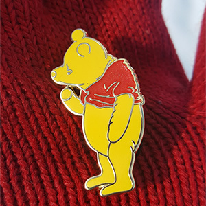 Modern photograph of an enamel pin in the shape of a teddy bear wearing a red shirt.