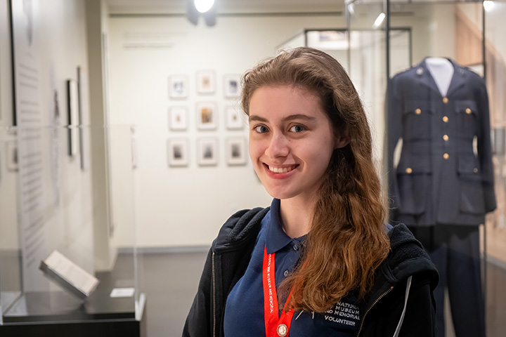 Modern photo of a smiling young white woman with long brown hair dressed in a blue polo shirt wearing a red lanyard, standing in a museum gallery.