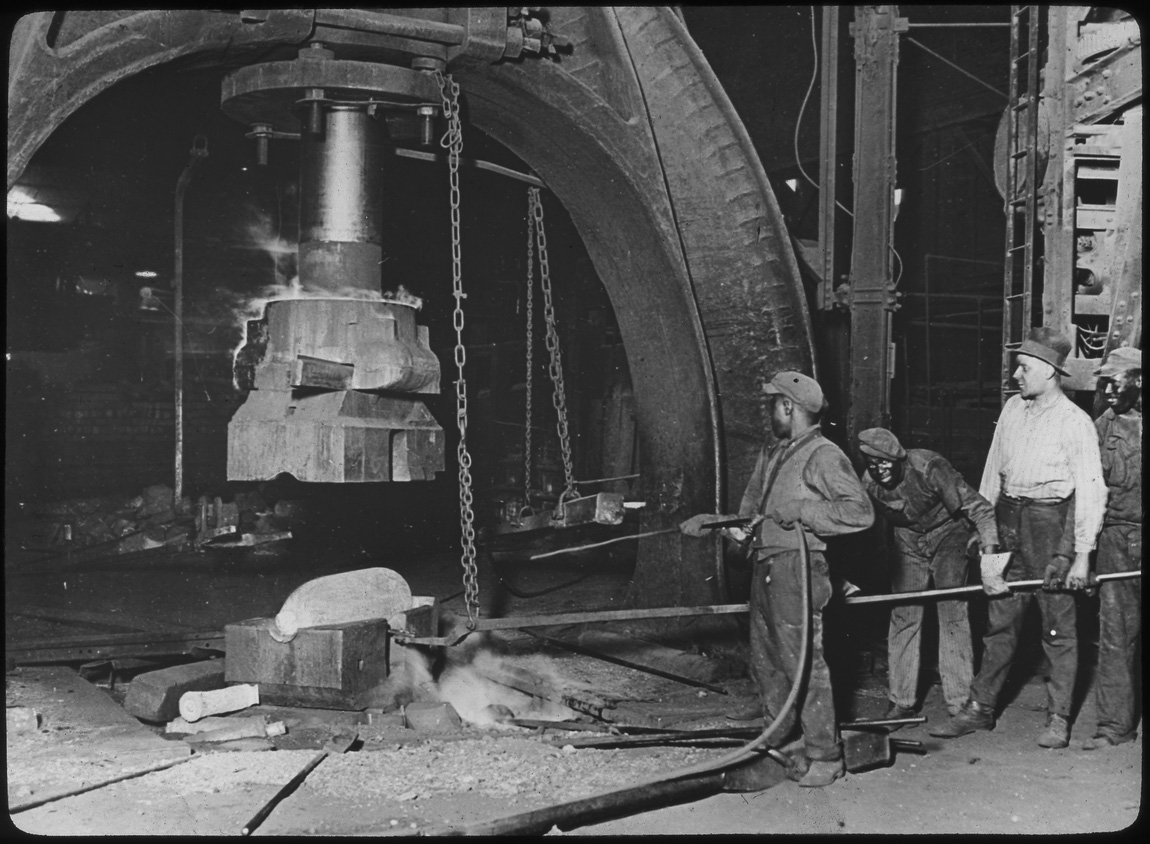 Black and white photograph of men forging large artillery shells in a factory.