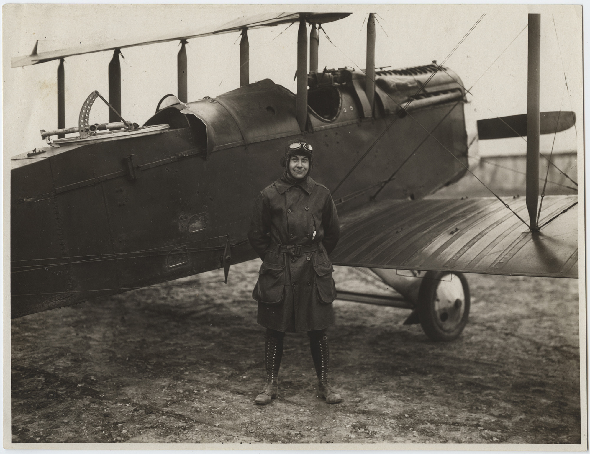 Black and white photograph of a man in a trench coat, tall lace-up boots, leather cap and goggles standing in front of a WWI biplane