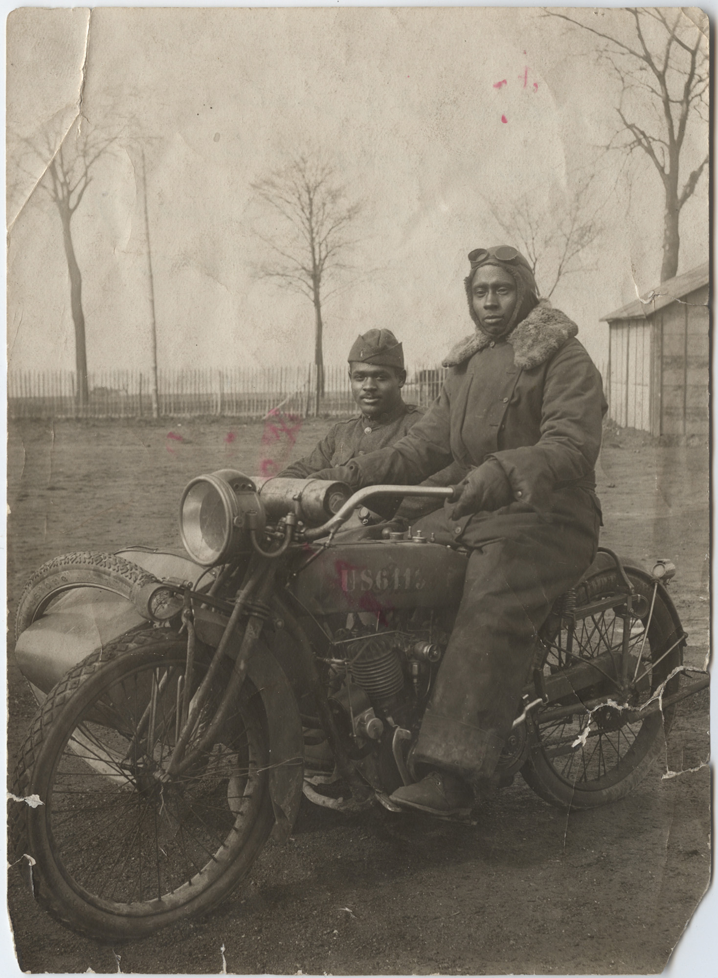 Black and white photograph of a Black man in a thick jumpsuit with a fur collar, a leather cap and goggles sitting on a motorcyle. Another Black man in ordinary uniform sits in the sidecar.