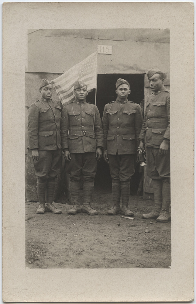 Black and white postcard photograph of four African American soldiers posing. One has an American flag tucked into his belt. Soldier on right is holding a bugle in his right hand.