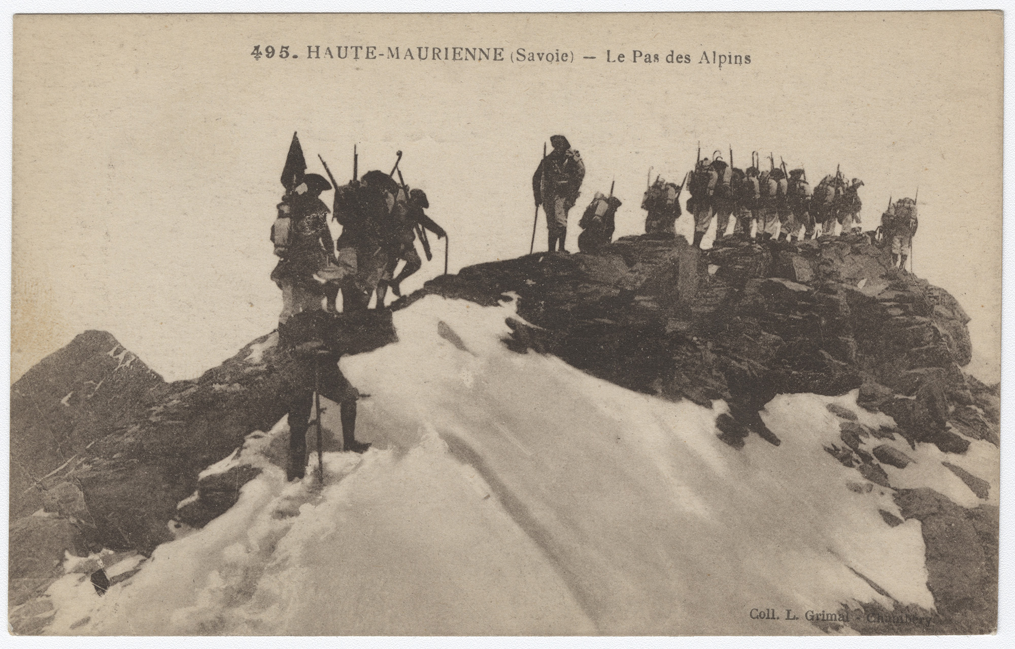 Black and white photograph of a line of soldiers weighed down with packs and weapons trudging over a mountain ridge