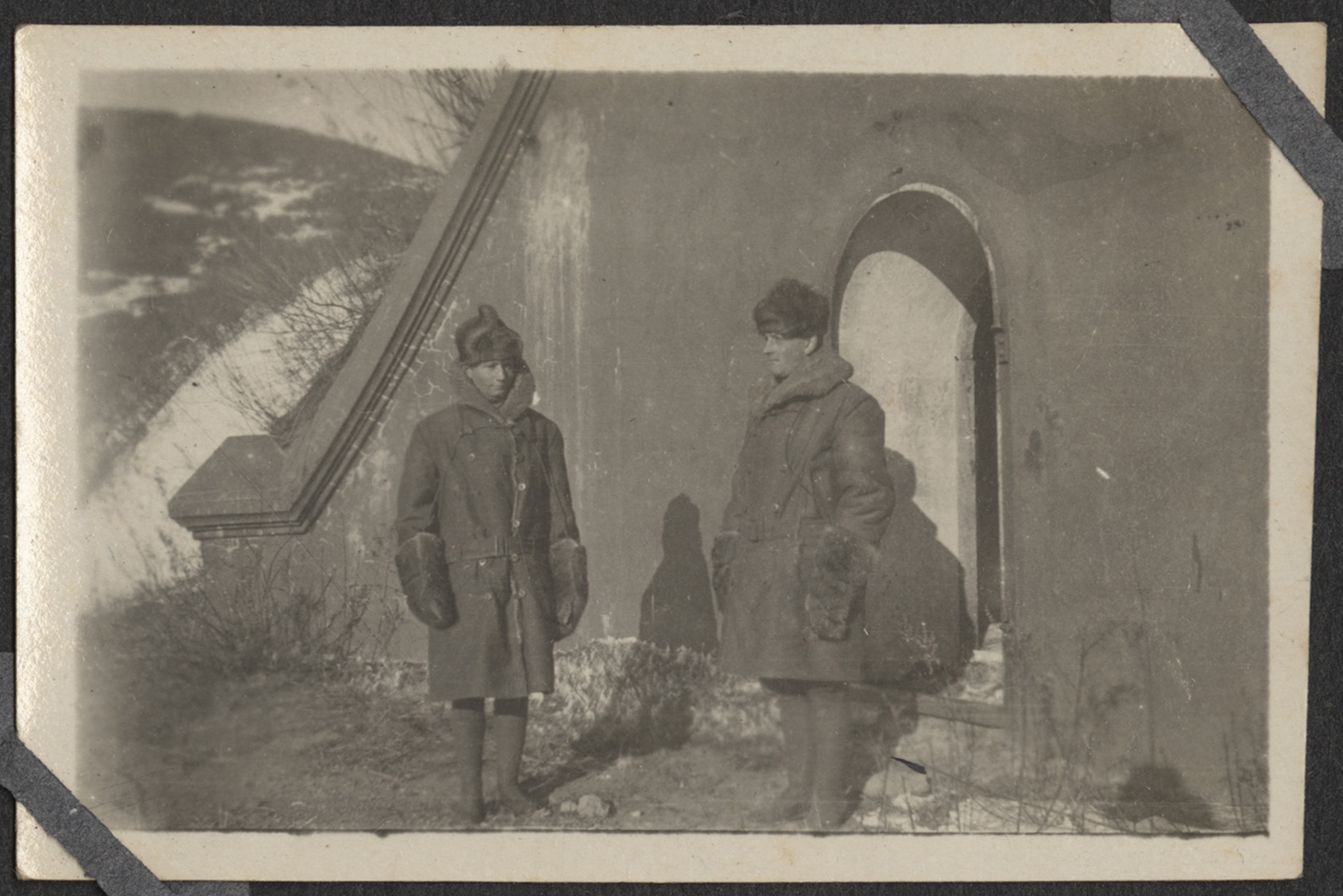 Black and white photograph of two men wearing thick coats, hats, boots and gloves standing outside in winter sunlight