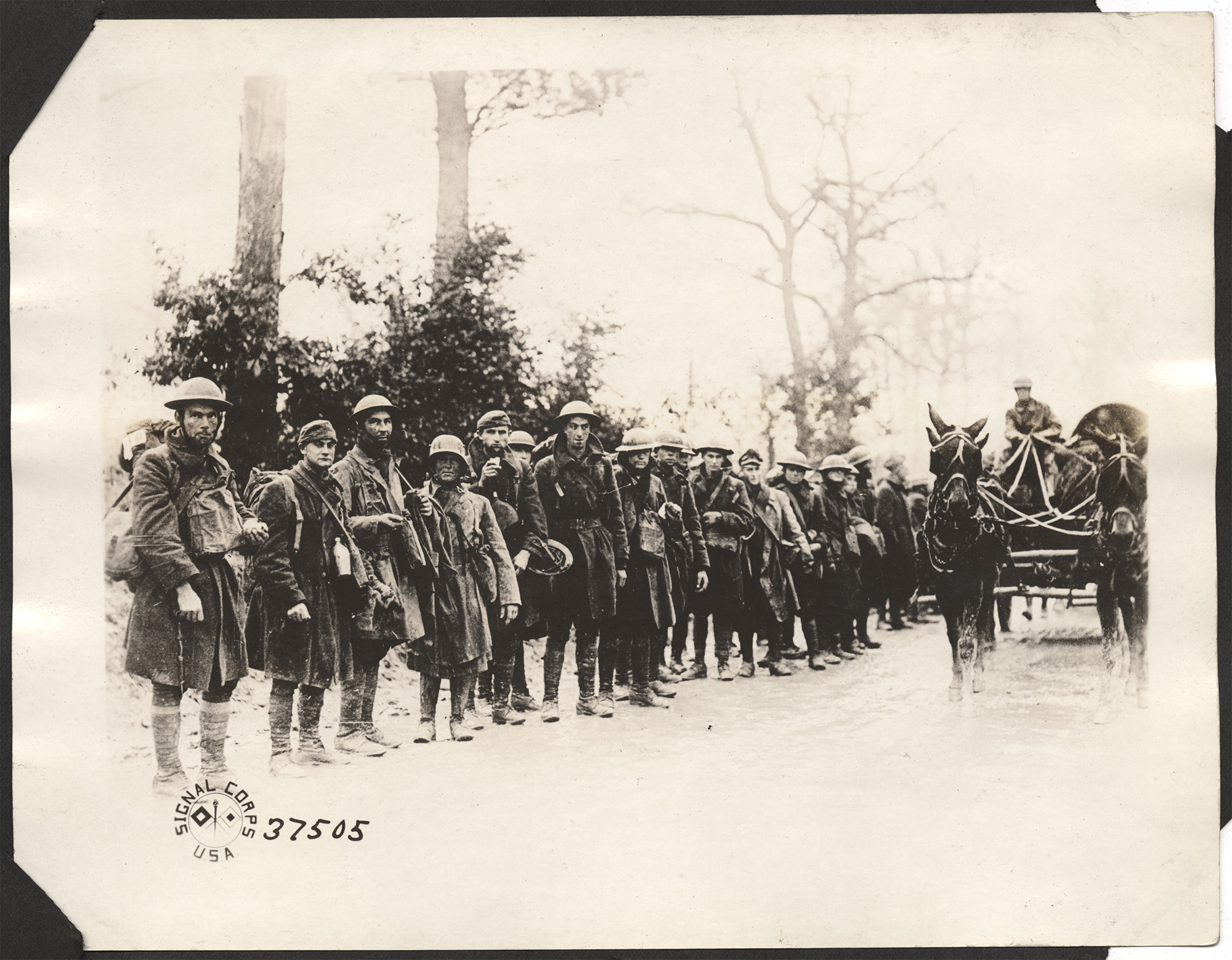 Black and white photograph of a large group of soldiers lined up next to a road and a horse-drawn cart. They are wearing rain coats and hats.