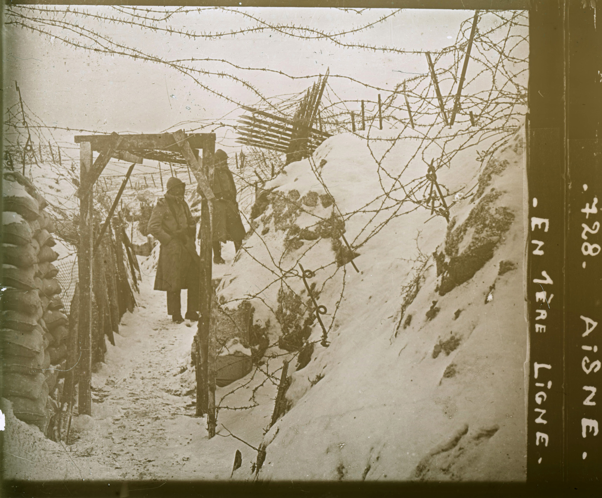 Black and white photograph of a trench covered in snow. A person in a long coat stands in the trench under scaffolding and barbed wire.