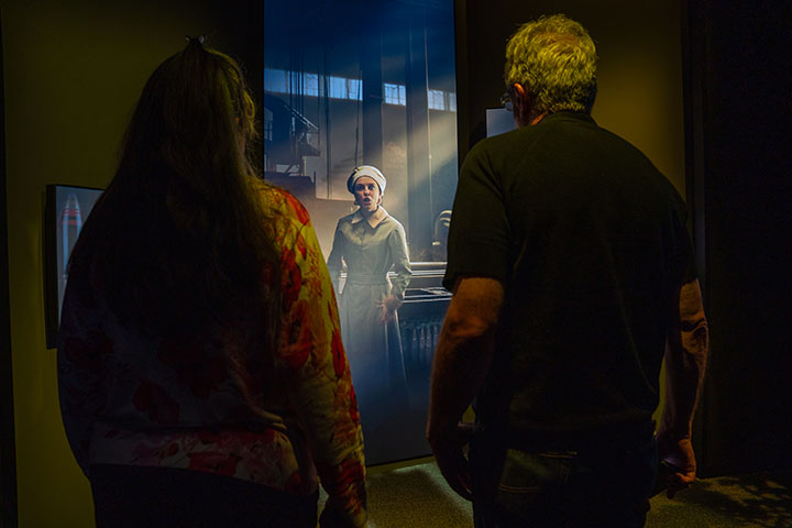 Guests watching a large video screen depicting a young white woman in a plain workday dress and cap standing on a factory floor