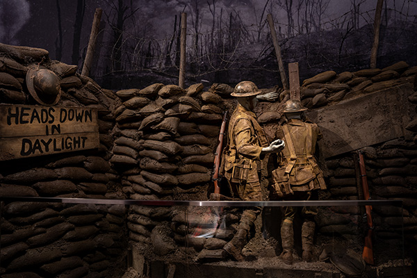 Museum exhibit replicating a British WWI trench with sandbags and wooden walkways. A soldier mannequin peers through a trench periscope while another soldier mannequin readies to throw a grenade.