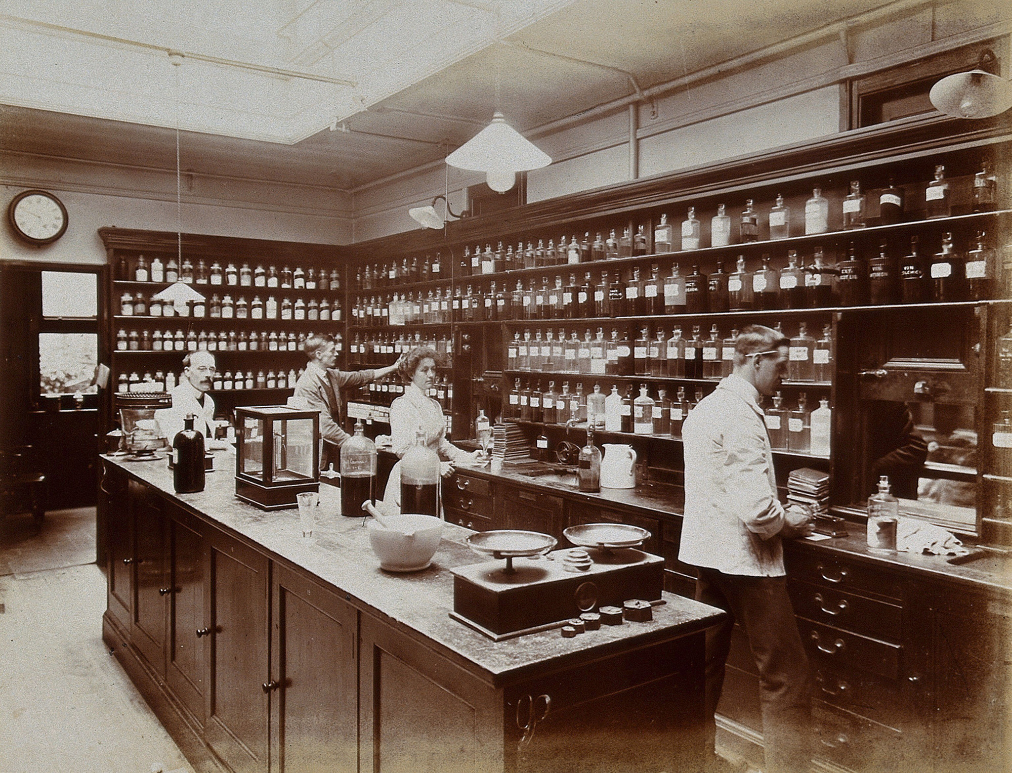 Black and white photograph of a pharmacy lined with dark wood shelves filled with jars. Four people are working behind the counter.