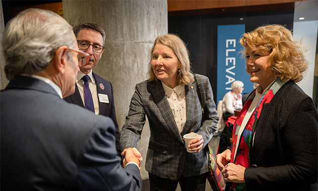 A blonde woman in a grey suit shakes hands with an older white man while two other people look on