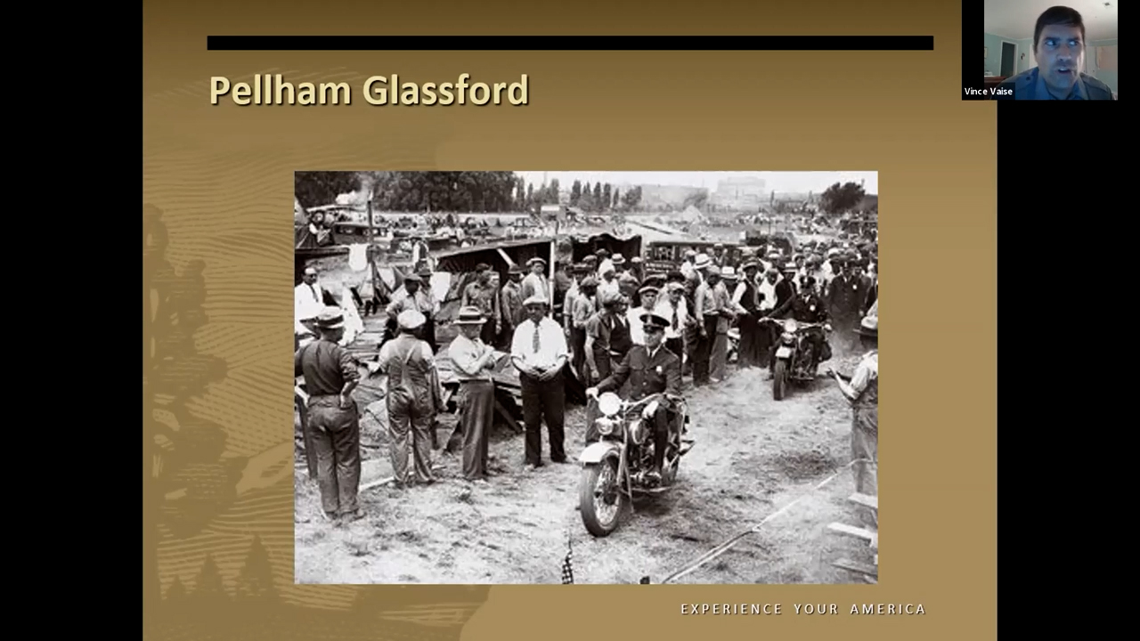 Black and white photo of crowds of men in an encampment. Through the middle, a man in a police uniform is riding a motorcycle